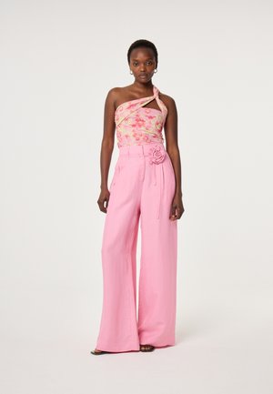 Vrouw die een roze asymmetrische bloemen top draagt en hooggetailleerde roze wijde broek met een stoffen bloemenriem.