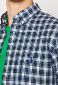 Košile s knoflíky v modro-zelené a bílé kostkované vzoru, vyrobená z lehkého bavlněného materiálu. Obsahuje logo na hrudi a zelené tričko pod ním.