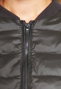 Svart puffjacka med horisontellt kviltat mönster. Har dragkedja fram och ribbad krage. Slät, glänsande tygtextur.