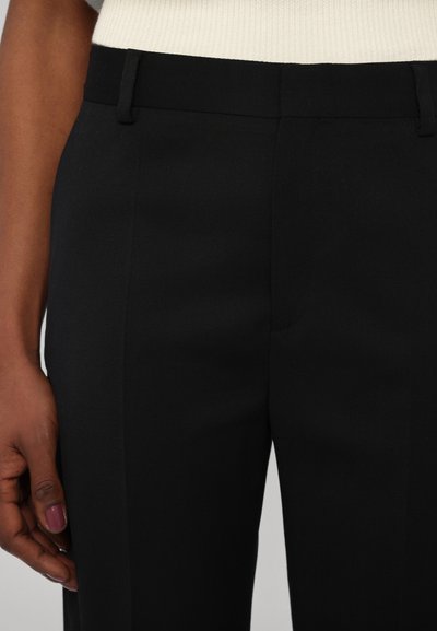 Zwarte broek met een gladde textuur, voorzien van een rechte voorkant, riemlussen en een slim fit. Hand houdt de zijkant vast, waarbij de nette naden zichtbaar zijn.