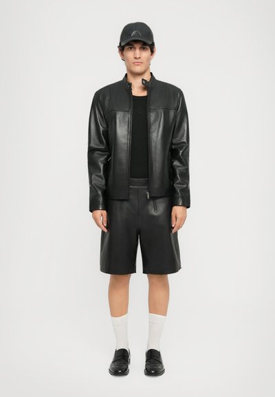 Man wearing black leather jacket and shorts, black shirt, black cap, white socks, and black loafers, standing against a plain background.