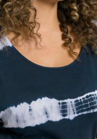 Lockigt hår person som bär en mörkblå skjorta med ett horisontellt vitt tie-dye-streck över bröstet.