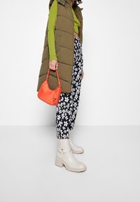 Olivově zelená nadýchaná vesta, dlouhé černé kalhoty s květinovým vzorem, světlé béžové kotníkové boty s masivní podrážkou a jasně oranžová kabelka.