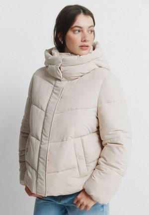 Kobieta ubrana w jasnobeżową, puchową kurtkę z wysokim kołnierzem, patrząca na bok, z rękami w kieszeniach, na jednolitym białym tle.