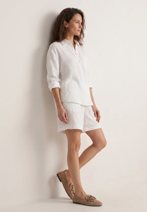 Vrouw in een witte gestructureerde blouse met knopen en bijpassende shorts, staand tegen een lichte muur, draagt beige loafers met kleurrijke kraalaccenten.