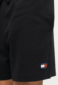 Κοντινό πλάνο μαύρων υφασμάτινων σορτς με μικρό έμβλημα Tommy Jeans κοντά στο τελείωμα στο δεξί πόδι.