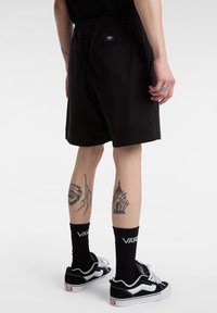 Pantaloni corti neri con cintura elastica, dotati di una tasca sul retro e un'etichetta con logo, abbinati a calze nere e sneakers nere.
