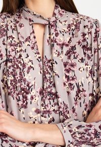 Květinová blůza z světle šedé látky, s malými fialovými a krémovými květy, s detaily na zavázání v oblasti krku, dlouhými rukávy a jemným leskem.