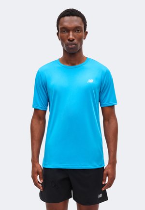 Hombre con camiseta deportiva de manga corta de color azul brillante de New Balance y pantalones cortos negros de New Balance, de pie frente a un fondo sencillo.