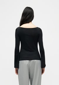 Γυναίκα με μακριά μαύρα μαλλιά, φορώντας μαύρη μπλούζα με μακριά μανίκια και γκρι ανοιχτό παντελόνι με ρίγες, εμφανίζεται από την πίσω όψη μπροστά σε απλό φόντο.