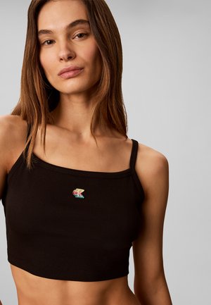 Ung kvinde med langt brunt hår iført en sort cropped tanktop med et lille flerfarvet "ck" logo.