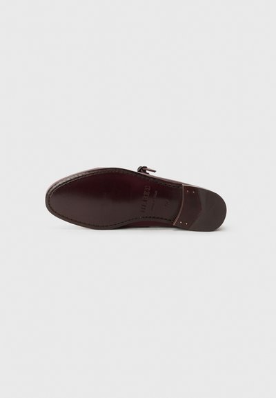 Suela de zapato de cuero, color burdeos oscuro, textura suave, forma redondeada, con herrajes minimalistas y un detalle de costura a lo largo del borde.
