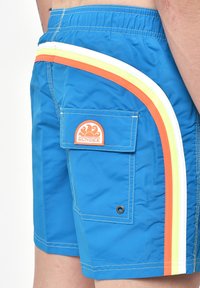 I pantaloni da bagno blu presentano una tasca laterale con un logo del sole arancione e tre strisce orizzontali gialle, arancioni e bianche. Tessuto morbido.