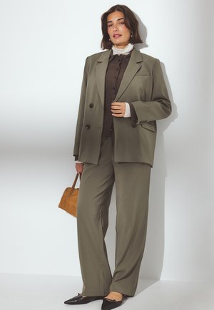 Donna che indossa un completo verde oliva con pantaloni larghi e un blazer over-size, camicia marrone, dolcevita bianca, scarpe flat nere e tiene in mano una borsa marrone.