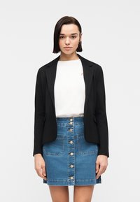 Jeune femme aux cheveux courts et foncés, portant un blazer noir, un t-shirt blanc et une jupe en jean boutonnée sur le devant, se tenant devant un fond blanc uni.