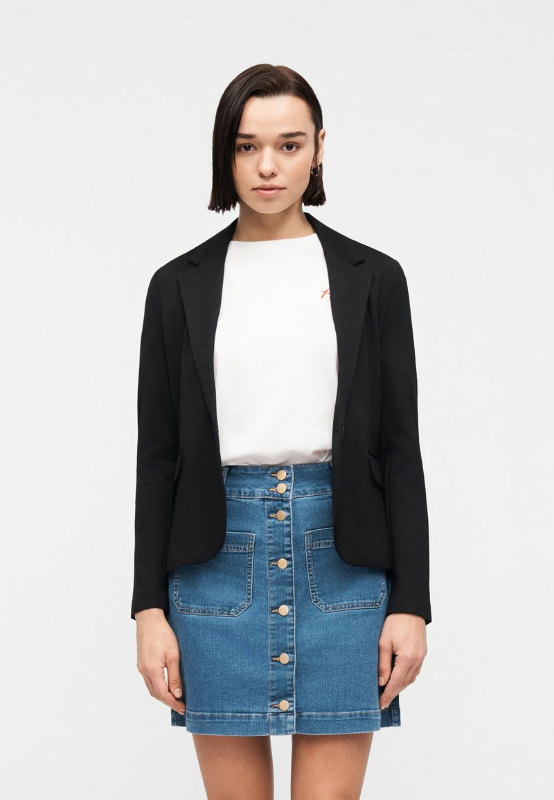 Jeune femme aux cheveux courts et foncés, portant un blazer noir, un t-shirt blanc et une jupe en jean boutonnée sur le devant, se tenant devant un fond blanc uni.