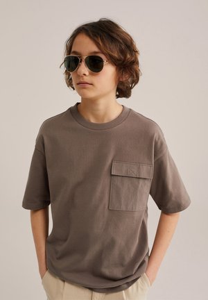 Jauns cilvēks ar īsiem brūniem matiem, valkā tumšas aviatora saulesbrilles, brūnu T-kreklu ar lielu priekšējo kabatu un gaišas krāsas bikses.