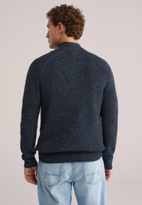 Tumši zils adīts džemperis ar teksturētu ribu rakstu, ar augstu apkakli un pielāgotām piedurknēm, saskaņots ar gaiši ziliem džinsa biksēm.