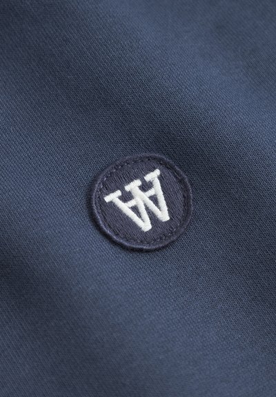 Tissu bleu marine au toucher lisse, arborant un patch brodé circulaire. Le patch affiche la lettre blanche "W" dans une police moderne et audacieuse.