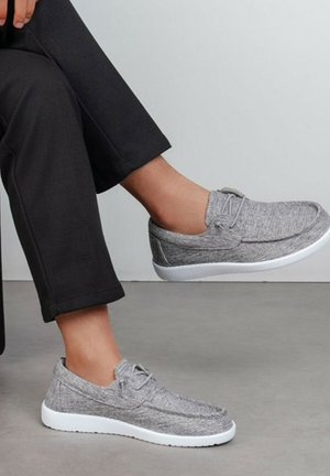 Personne portant des chaussures décontractées grises à enfiler avec des semelles blanches, assise avec une jambe croisée sur l'autre, portant un pantalon noir.