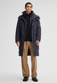 Mann in einem langen, dunkelblauen Puffy-Mantel über einem schwarzen Strickpullover, beigen weit geschnittenen Hosen und schwarzen Schuhen, der vor einem neutralen Hintergrund steht.
