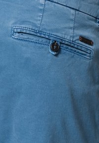 Blå bomullbyxor med en knäppt bakficka med sydd kant. En liten märkesetikett syns nära fickan.