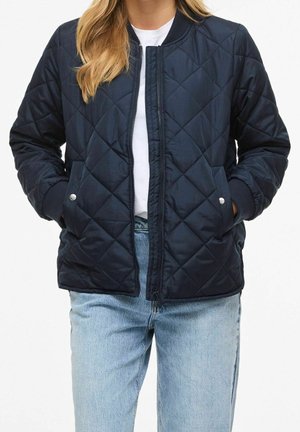Mujer con chaqueta bomber acolchada azul marino con bolsillos de botones de presión, camisa blanca y vaqueros azul claro, manos en los bolsillos de la chaqueta.