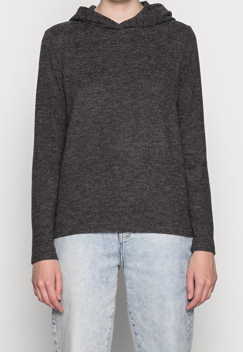 Personne portant un sweat à capuche gris foncé à manches longues et un jean bleu clair, se tenant devant un fond blanc uni.