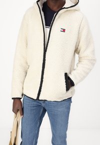 Man som bär en krämfärgad fleecejacka med svart dragkedja och fickor, blå jeans och håller en beige tote-väska med ena handen.