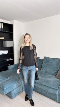 Črna dolga majica z prosojnimi cvetličnimi čipkastimi rokavi, v kombinaciji s svetlo modrimi kavbojkami in črnimi loaferji, drži majhno črno torbico.