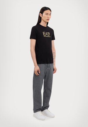 Camiseta de algodón negra con texto de logo en dorado, cuello redondo, mangas cortas, combinada con jeans holgados grises y zapatillas blancas.