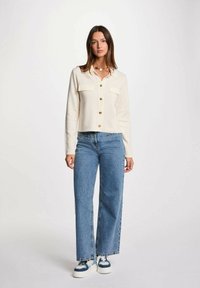 Cardigan crema con quattro bottoni dorati e due tasche sul petto, abbinato a jeans a gamba larga blu e sneakers bianche con dettagli blu.