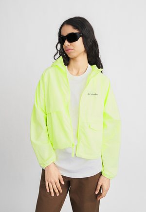 Γυναίκα που φοράει μαύρα γυαλιά ηλίου, φωτεινό κίτρινο μπουφάν Columbia με φερμουάρ, λευκό μπλουζάκι και καφέ παντελόνι, στέκεται μπροστά σε ανοιχτό γκρι φόντο.