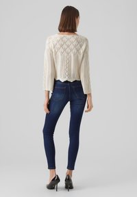 Maglione lavorato a maglia crema con un motivo geometrico e maniche a tre quarti, abbinato a jeans skinny blu scuro e scarpe con tacco nere.