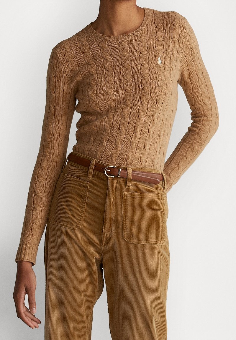 Femme portant un pull beige en maille torsadée avec petit logo, pantalon en velours marron et ceinture en cuir marron sur fond blanc uni.