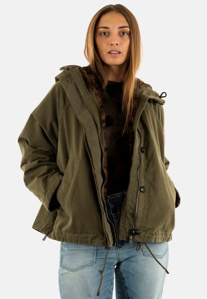 Femme aux longs cheveux bruns portant une veste olive ouverte avec des cordons, un haut noir et un jean bleu clair, se tenant contre un fond blanc.