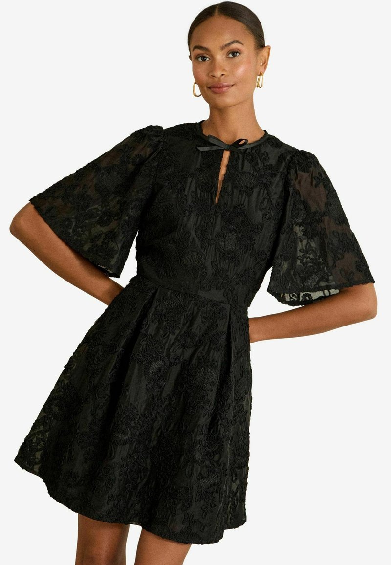 Juoda nėriniuota suknelė su pločiais trumpais rankovėmis, gėlių siuvinėjimu, raktų skylutės iškirpte ir prigludusia talija, pereinančia į plačią sijoną.