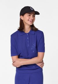 Lila Poloshirt mit kurzen Ärmeln und strukturierter Oberfläche, das mit einem floralen Stickdesign und einem Polokragen versehen ist. Kombiniert mit einer passend gefärbten Hose.