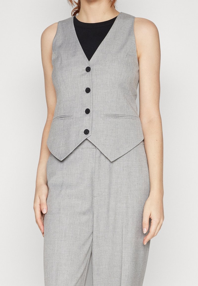 Femme portant un gilet gris clair boutonné sur un haut noir avec un pantalon gris assorti, debout devant un fond uni.