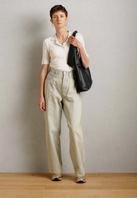 Personne portant un haut blanc à manches courtes, un pantalon beige taille haute à jambes larges, des baskets dorées et tenant un sac noir sur l'épaule.