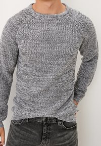 Grau gemusterter Strickpullover mit Rundhalsausschnitt und langen Raglanärmeln, der aus einem strukturierten, weichen Stoff besteht und zu dunklen Jeans passt.