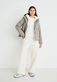 Jachetă gri tip puffer cu guler pufoș, pulover alb texturat dedesubt, pantaloni largi crema și adidași gri cu accente negre.