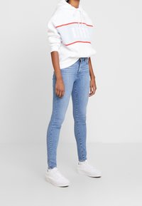 Felpa bianca con una banda orizzontale blu e rossa che riporta la parola "LIMITS", abbinata a jeans skinny azzurri e sneakers bianche.