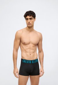 Black boxer briefs with a thick, turquoise "CALVIN KLEIN" logo waistband. The fabric appears smooth and form-fitting. Model shown is shirtless.