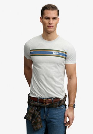 Man draagt een lichtgrijs aansluitend T-shirt met blauwe, groene en oranje strepen over de borst, spijkerbroek, bruine riem en een geruit overhemd gestrikt om de taille.