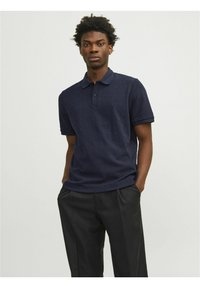 Polo azul marino con un patrón de diamante texturizado, tapeta de tres botones y mangas cortas. Combinado con pantalones de vestir negros.