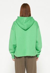 Sudadera verde hecha de tejido texturizado, con capucha, corte holgado y dobladillo ligeramente recortado. El modelo lleva pantalones marrones de corte holgado.