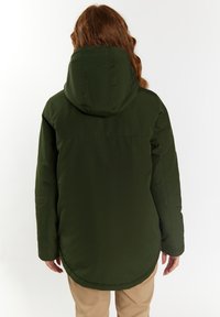 Chaqueta aislante verde con capucha, de textura suave y con dobladillo redondeado, llevada sobre pantalones de colores claros, vista desde la parte de atrás.