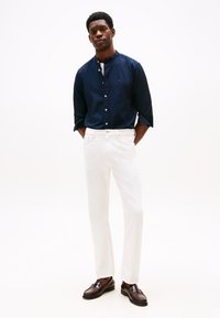 Camisa azul marino con botones y cuello mao, pantalones vaqueros blancos de pierna recta y mocasines marrones, posando contra un fondo blanco liso.