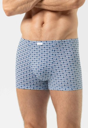 Man draagt blauwe boxershorts met kleine geometrische patronen, staat met gekruiste armen tegen een effen achtergrond.